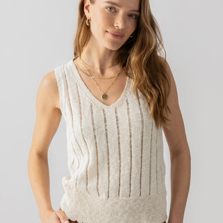 A woman with long, light brown hair is wearing the "Vacation Mode Shell Birch" by Sanctuary Clothing along with brown pants. She has her hands in her pockets, and she is adorned with gold hoop earrings and two necklaces. She stands against a plain, light-colored background and smiles slightly.