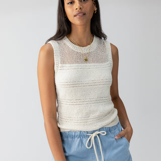 A person with long dark hair is wearing Sanctuary Clothing's Waves Of Summer Shell Birch, a sleeveless cream-colored knitted top, paired with light blue drawstring shorts. They are posed with one hand in their pocket against a plain, light-colored background and accessorized with a gold necklace and hoop earrings.