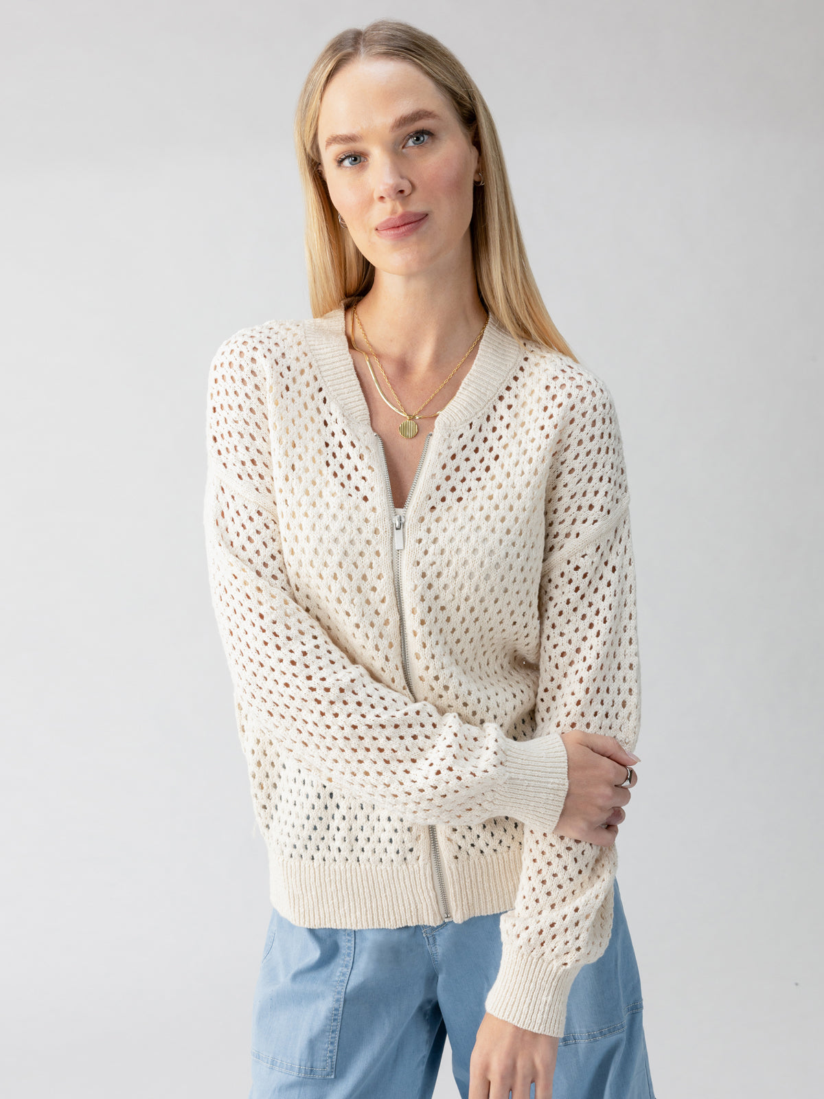 A woman with long blonde hair is wearing a cream-colored Stepping Out Bomber Eco Natural from Sanctuary Clothing over a light-colored top and light blue pants. She has a neutral expression and is looking directly at the camera, against a plain, light-colored background.