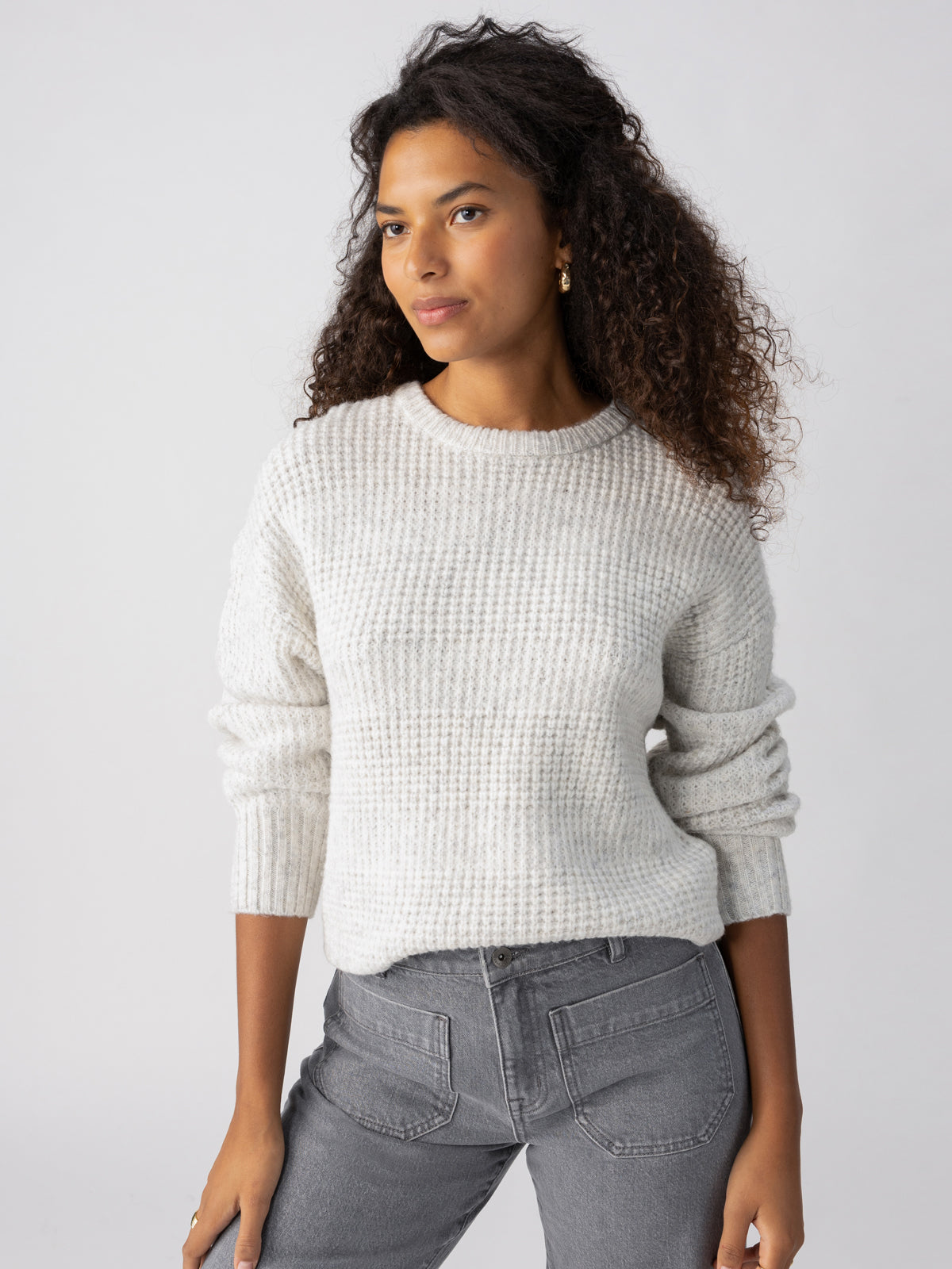 A person with long curly hair wearing a Sanctuary Clothing Snow Bunny Sweater Chalk Heather and gray jeans poses against a plain white background. They have a confident expression, with one hand resting on their hip and the other arm relaxed by their side.