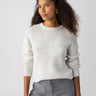 A person with long curly hair wearing a Sanctuary Clothing Snow Bunny Sweater Chalk Heather and gray jeans poses against a plain white background. They have a confident expression, with one hand resting on their hip and the other arm relaxed by their side.