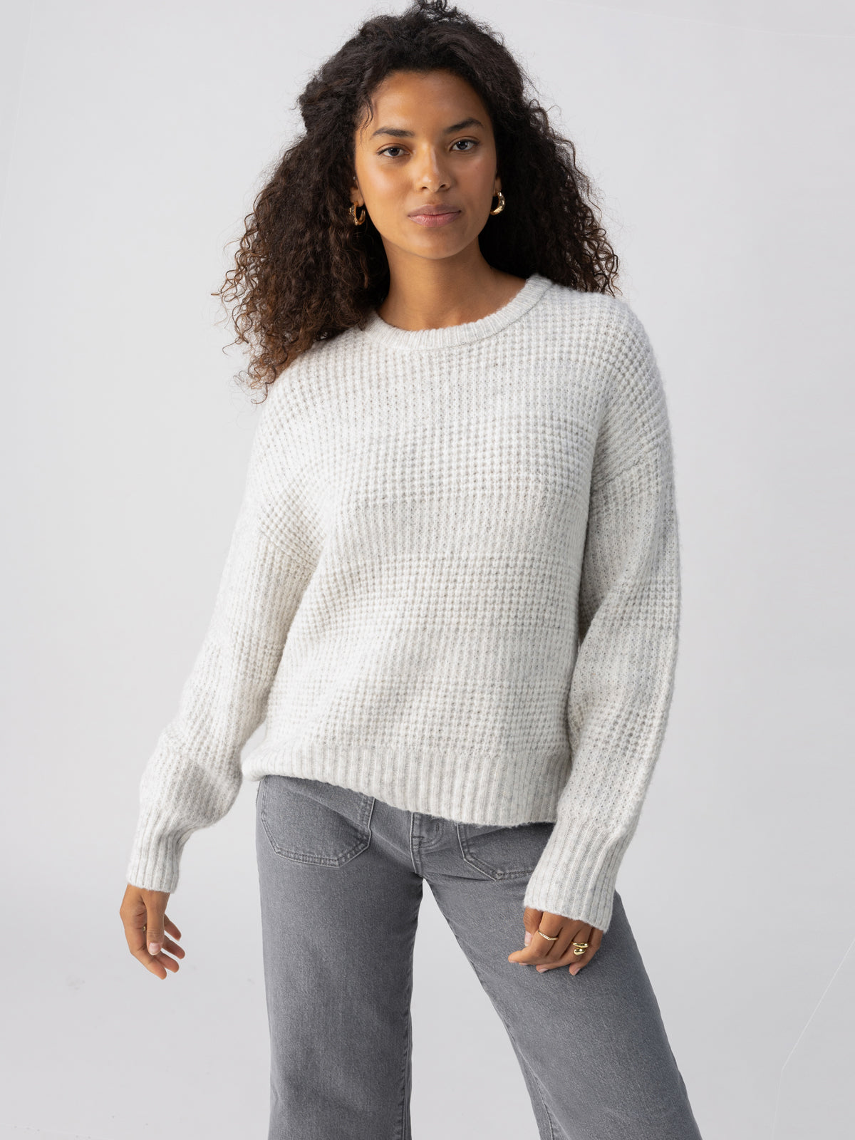 A person with long, curly hair is wearing the cozy, oversized "Snow Bunny Sweater Chalk Heather" by Sanctuary Clothing, paired with gray jeans. They stand against a plain light background with a relaxed expression and one arm slightly bent.