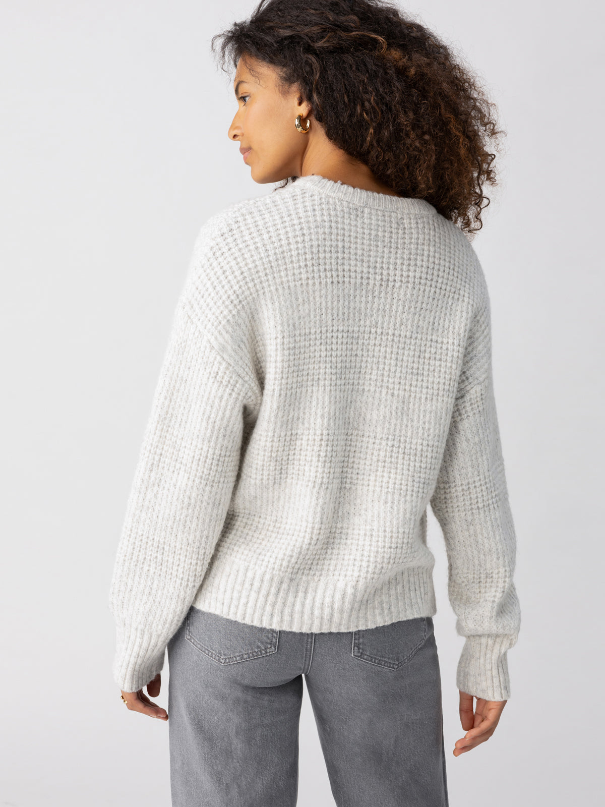 A person with curly hair is wearing a Sanctuary Clothing Snow Bunny Sweater in Chalk Heather and gray jeans. They are standing with their back to the camera, looking slightly to the side against a plain white background.