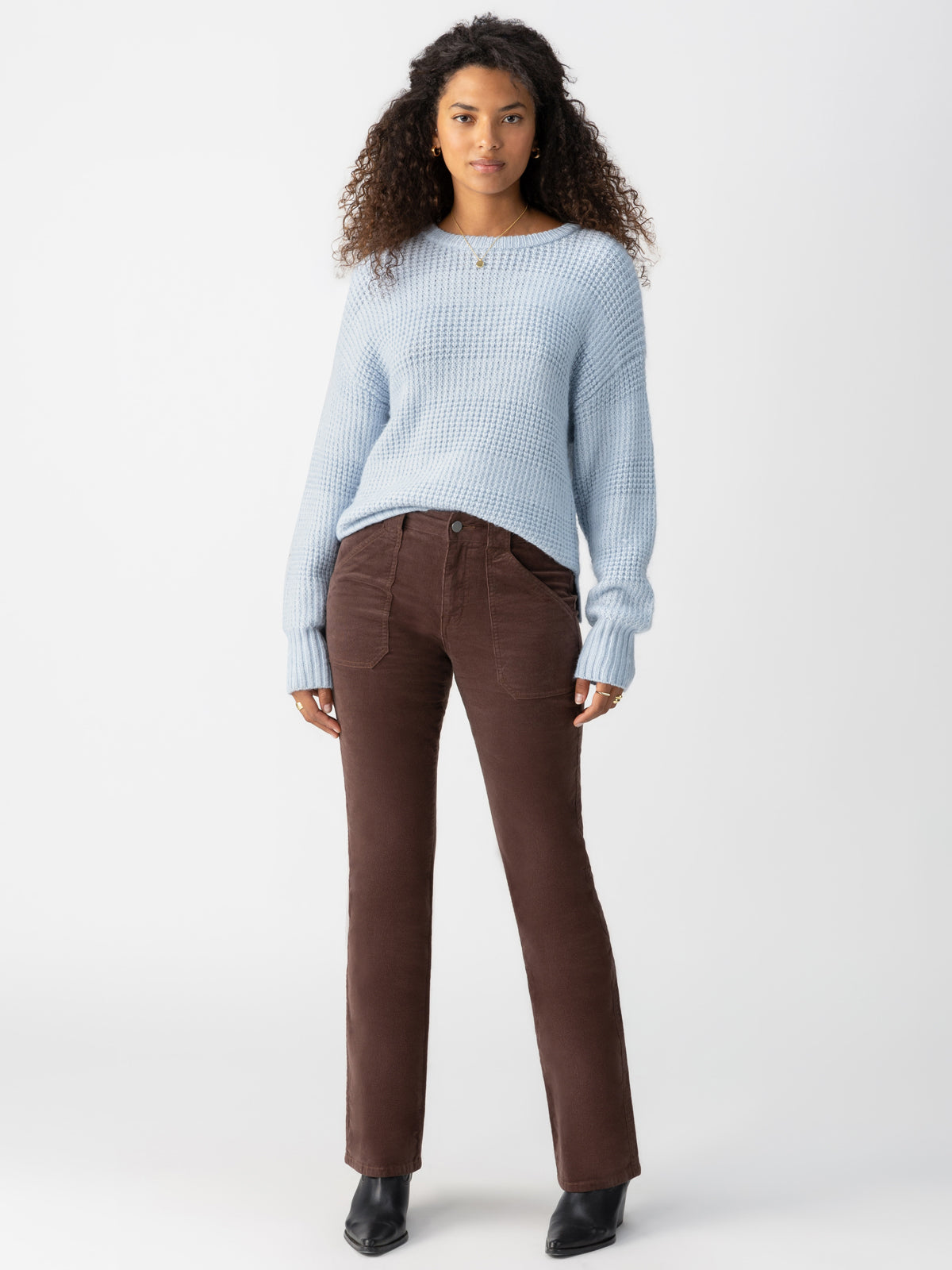 A woman with curly hair is standing against a plain background, wearing the Snow Bunny Sweater Sky by Sanctuary Clothing, paired with brown corduroy pants and black ankle boots. Her arms are relaxed by her sides and she is facing the camera.