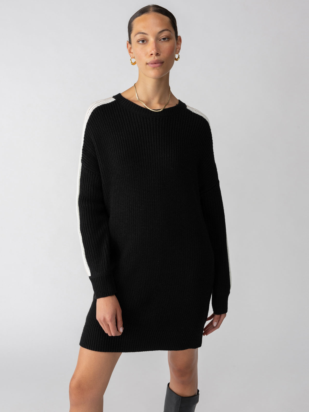 A person stands against a plain background, wearing the Speedway Sweater Dress Black Milk Toasted Almond by Sanctuary Clothing. They have pulled-back hair, hoop earrings, and boots. Their arms are relaxed by their sides.