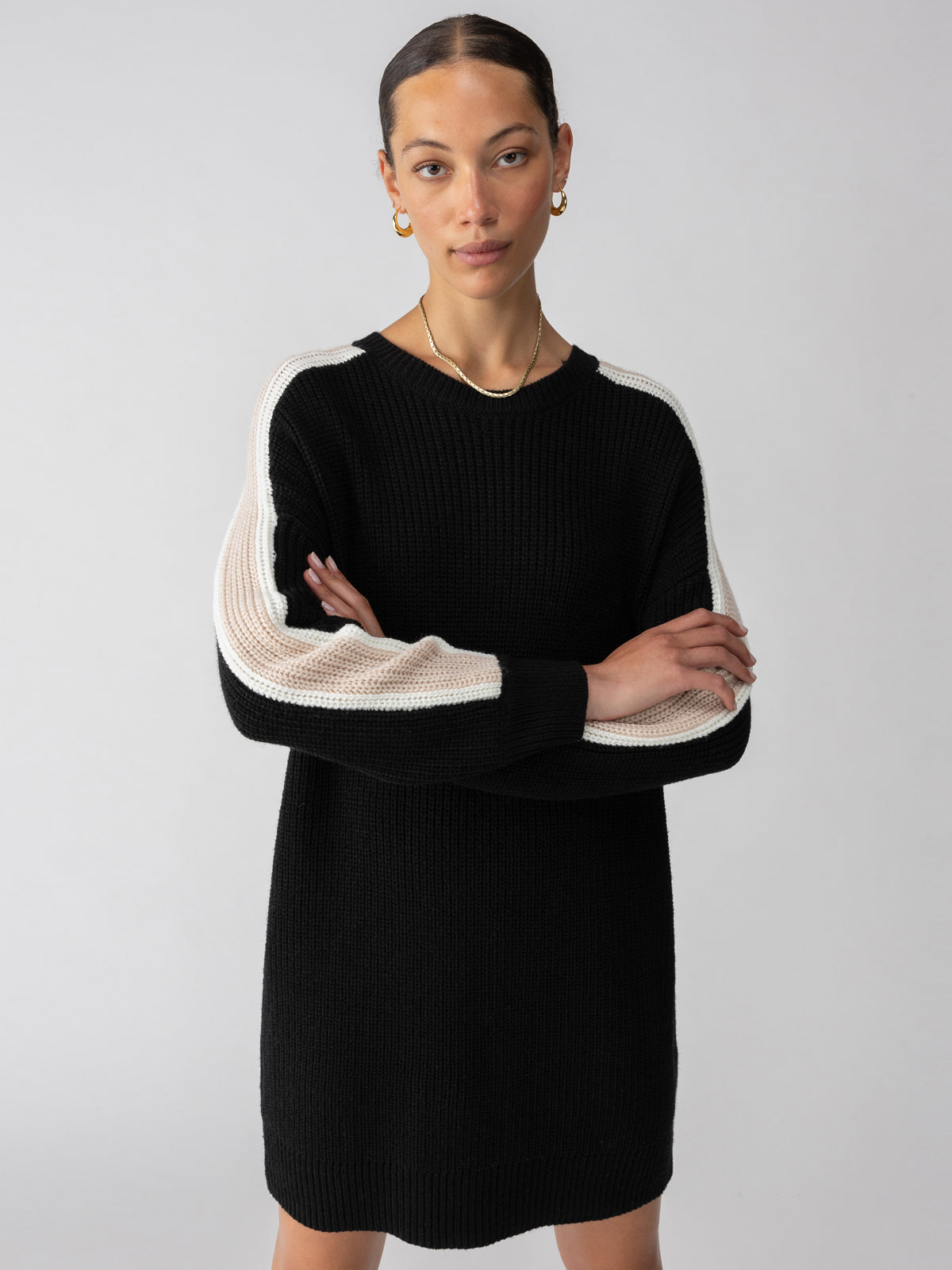 A person wearing the Speedway Sweater Dress Black Milk Toasted Almond by Sanctuary Clothing, featuring white stripes along the shoulders and outer arms, stands against a plain, light grey background. They have their arms crossed and are looking directly at the camera while sporting gold hoop earrings.