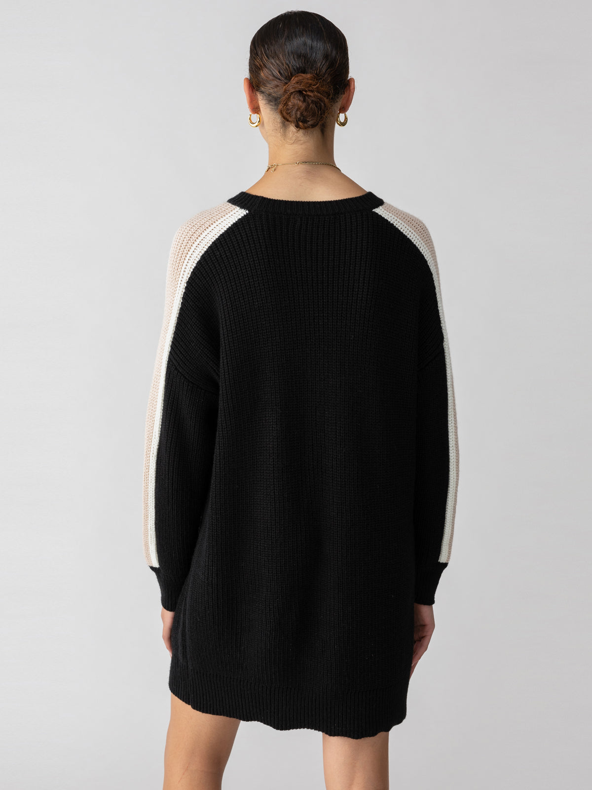 A person with their hair tied back is seen from behind wearing the Sanctuary Clothing Speedway Sweater Dress in Black Milk Toasted Almond, accessorized with hoop earrings and a chain necklace. They are standing against a plain light gray background.