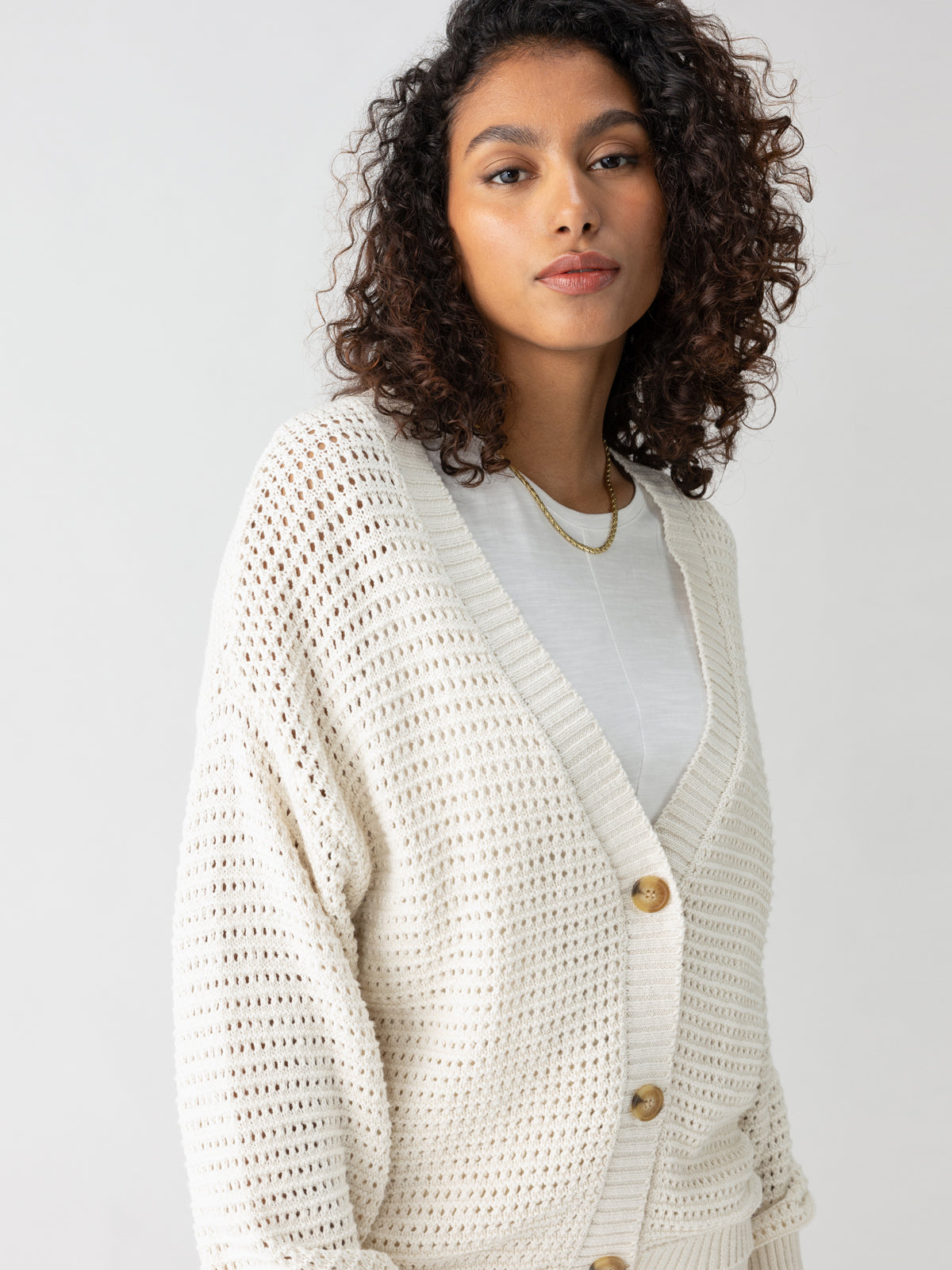 A woman with curly hair wears the Falling For Fall Cardi Chalk by Sanctuary Clothing over a white top. The off-white knitted cardigan features large buttons and a loose, textured pattern. She looks towards the camera with a neutral expression against a plain light background.