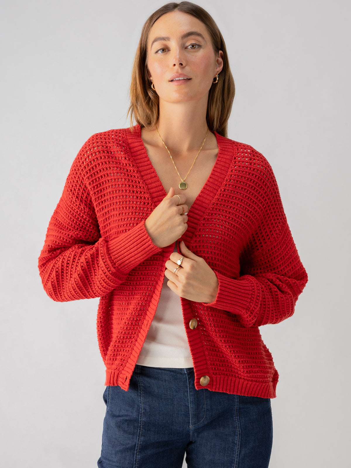 A person with long, light brown hair wears the Sanctuary Clothing Falling For Fall Cardi in Cherry Red over a white top. They have a calm expression, stand against a plain gray background, and hold part of the cardigan with one hand. They also wear a gold necklace.