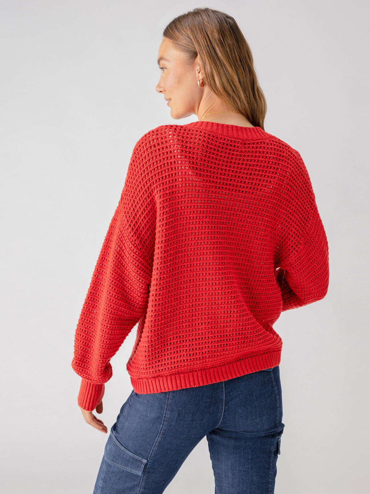 From behind, a person with long hair is shown wearing the Sanctuary Clothing "Falling For Fall Cardi Cherry Red," a bright red, textured knit sweater. Paired with fitted, dark blue jeans, the individual stands against a plain and light-colored background.