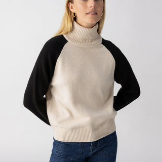 A person with long blonde hair is wearing a cream-colored Cozy Day Sweater in the Toasted Almond Black style by Sanctuary Clothing, paired with blue jeans. They have a neutral expression and are standing in front of a plain, light gray background. Their hands are behind their back.