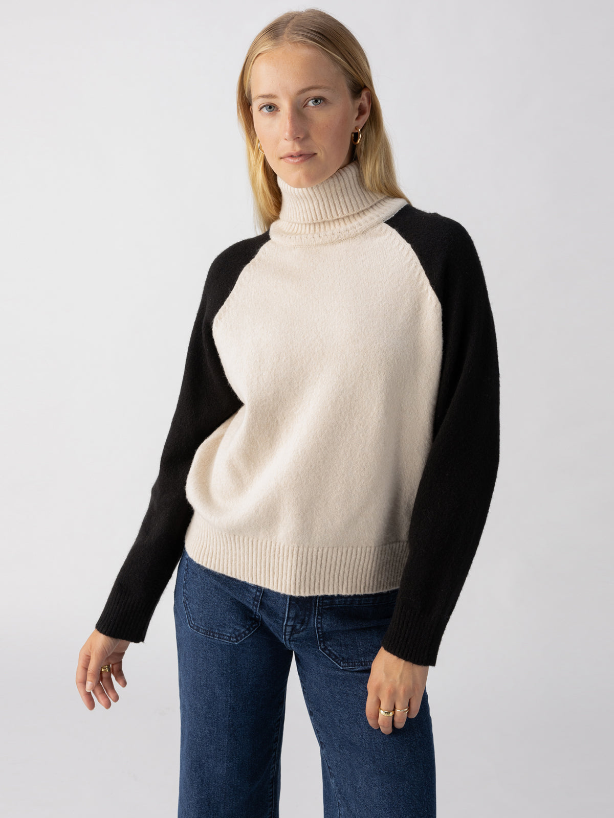 A woman with light hair stands against a plain background, wearing the Cozy Day Sweater in Toasted Almond Black by Sanctuary Clothing, paired with blue jeans. She is looking directly at the camera with a neutral expression.