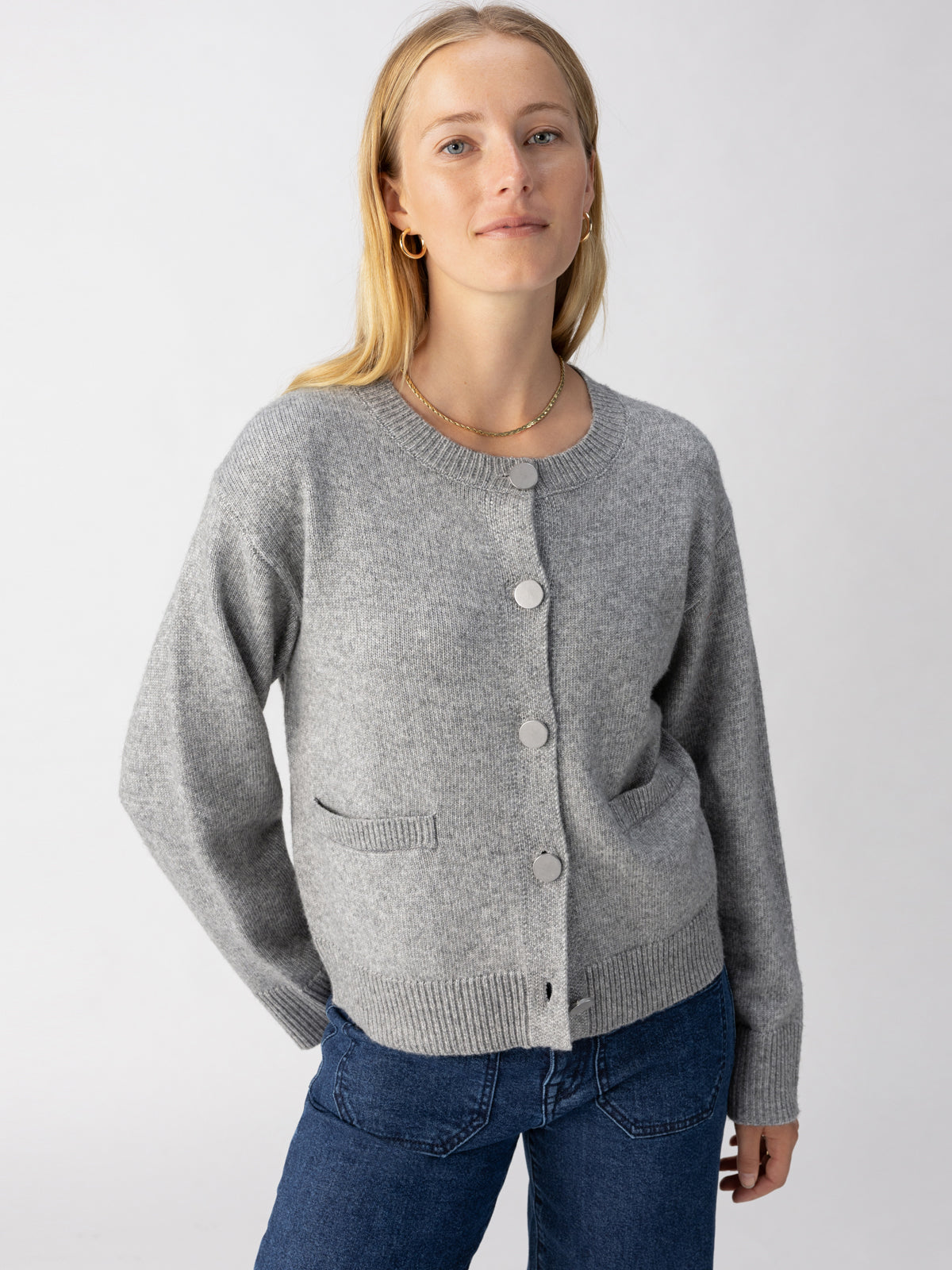 A person with long, blonde hair is standing against a plain background, wearing the Elevated Cardi Heather Ash by Sanctuary Clothing and blue jeans. They have a calm expression and their hands are resting by their sides.