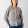 A person with long, blonde hair is standing against a plain background, wearing the Elevated Cardi Heather Ash by Sanctuary Clothing and blue jeans. They have a calm expression and their hands are resting by their sides.