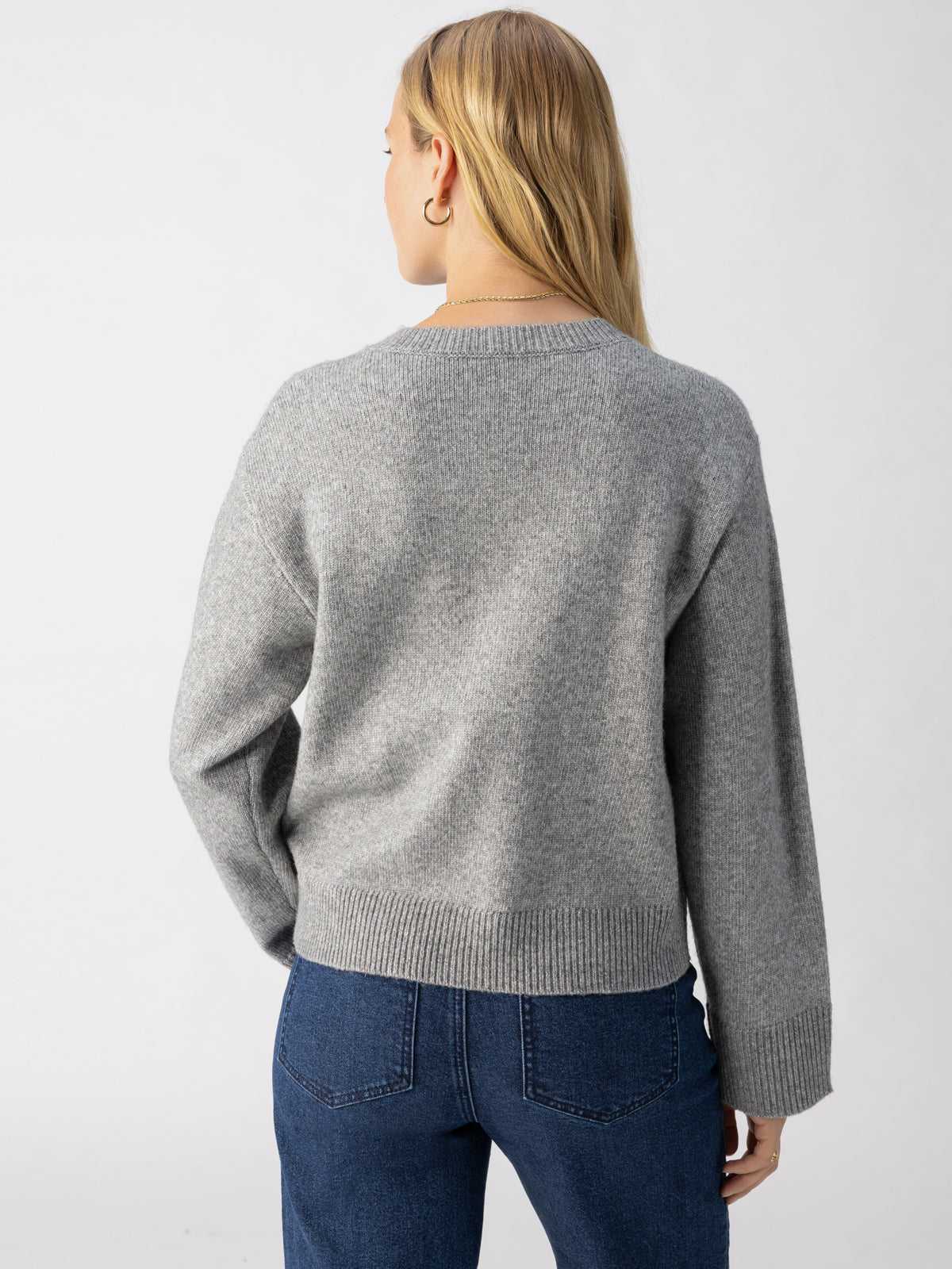 A person with long blonde hair, seen from the back, is wearing the Elevated Cardi Heather Ash by Sanctuary Clothing and blue jeans. The background is plain and light-colored.