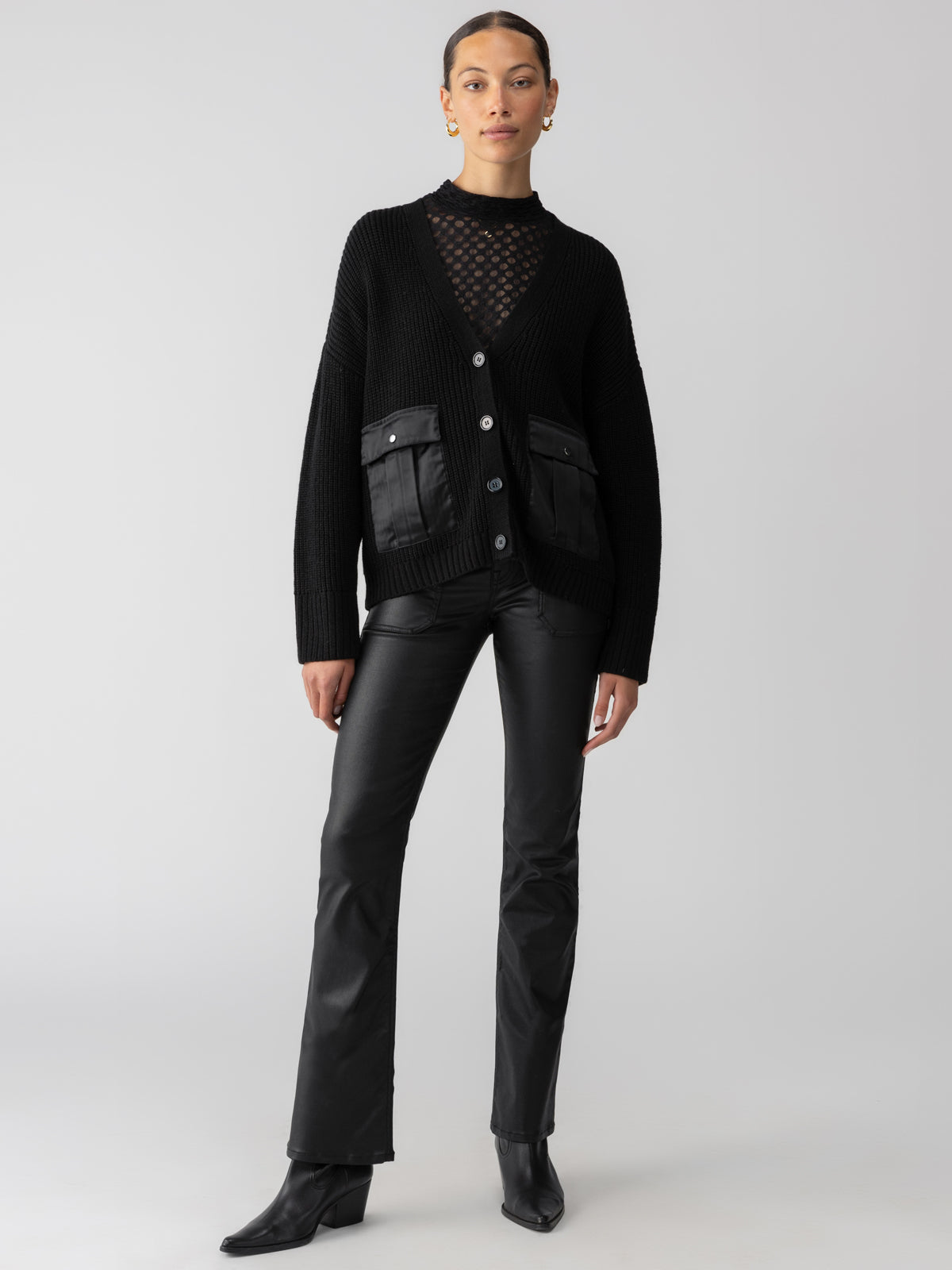 A person stands confidently, wearing the Sanctuary Clothing's "Trend Setter Cardi Black" over a sheer black top, paired with black pants and heeled ankle boots. The cardigan features two front pockets with leather detailing and large buttons. The background is a plain, light gray color.