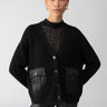 A person with hair pulled back and gold earrings wears the Trend Setter Cardi Black from Sanctuary Clothing, which features buttons and front pockets, layered over a black polka-dot top. They stand against a plain light gray background with hands resting on their hips.