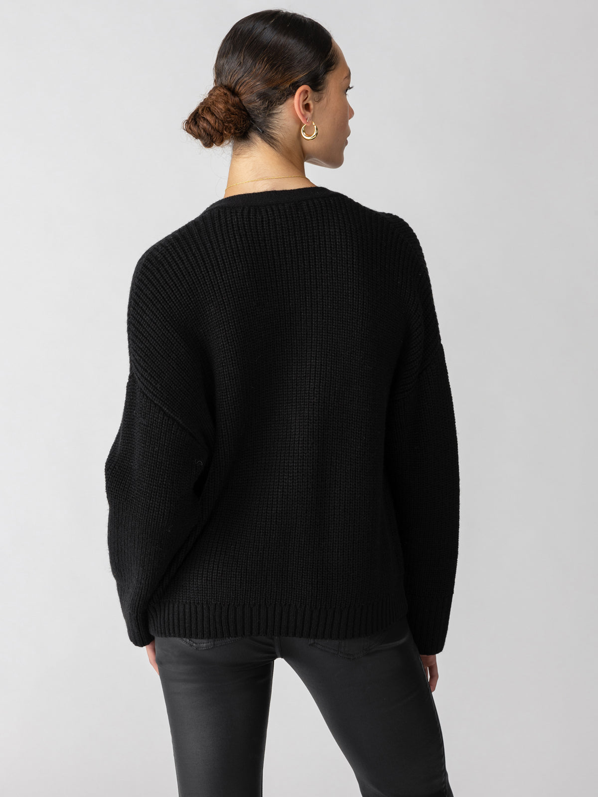 A person with tied-back hair wearing the Trend Setter Cardi Black from Sanctuary Clothing and black pants is shown from the back. The cardigan has a loose fit with a ribbed texture. The background is plain and light-colored.