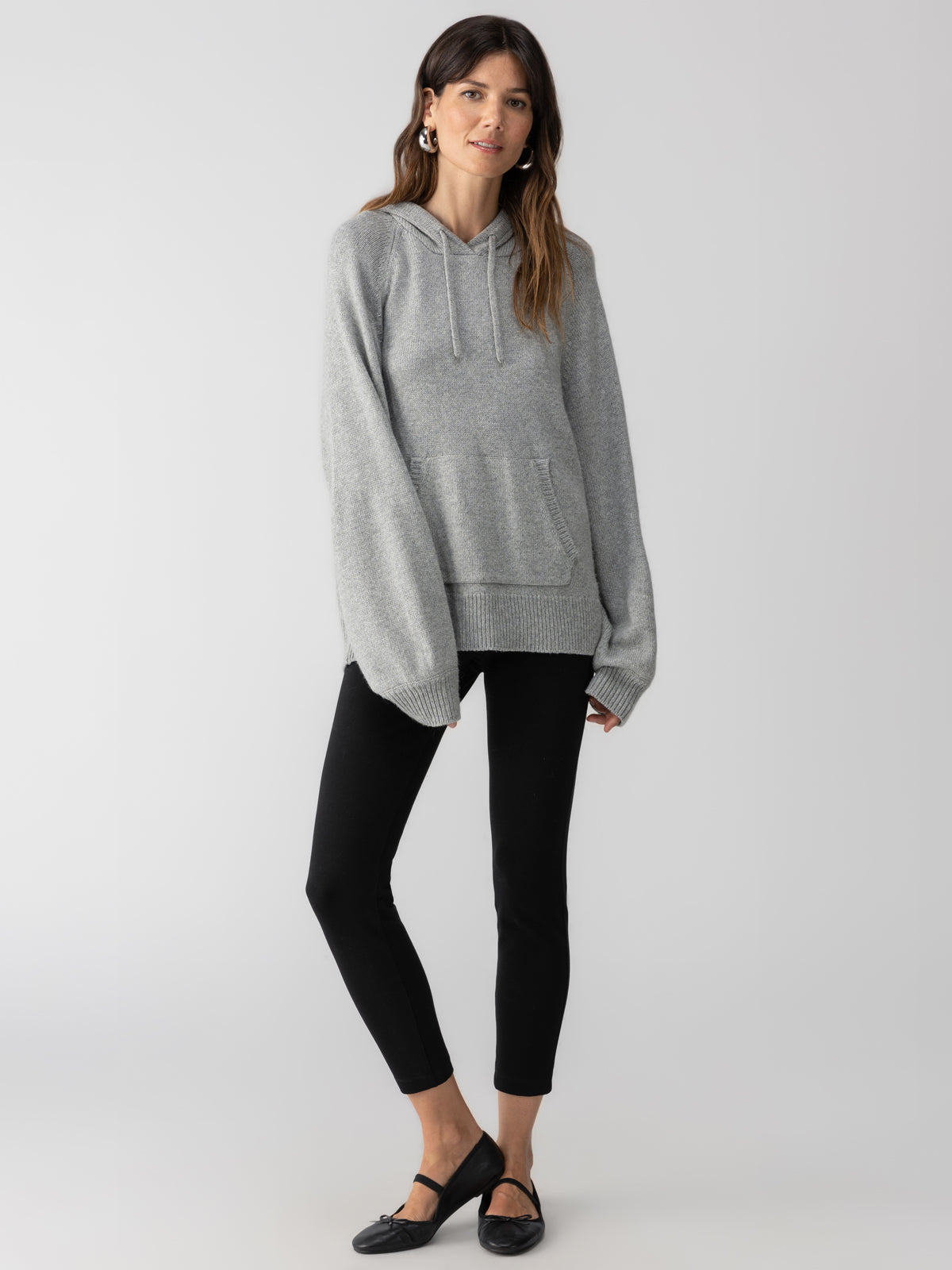 A person is standing against a plain background, wearing the Elevated Layering Hoodie in heather grey by Sanctuary Clothing, paired with black leggings and black flats. They have long hair and are posed with one leg slightly crossed over the other.