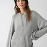 A woman with long brown hair is wearing the Sanctuary Clothing Elevated Layering Hoodie in Heather Grey and black pants. She stands against a plain white background, looking directly at the camera with a relaxed expression and her right hand on her hip.