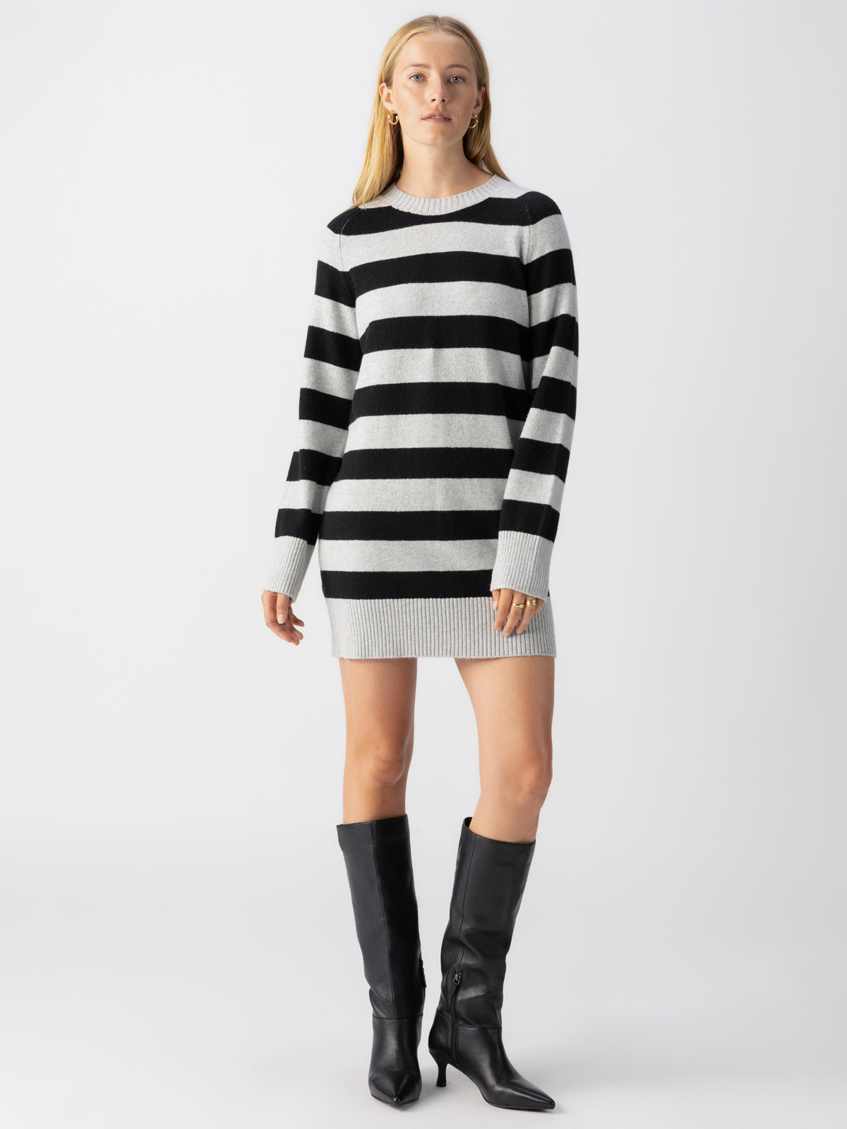 A person is posing in the Sanctuary Clothing So Soft Sweater Dress Heather Grey Black Stripe and black knee-high boots. They have long hair and are standing against a plain white background.