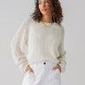 Someone with curly hair is dressed in the Perfect Moment Sweater Chalk by Sanctuary Clothing, featuring mesh sleeves, and paired with white high-waisted pants. Their hands are in their pockets, and they are gazing to the side with a neutral expression. The background is plain and light-colored.