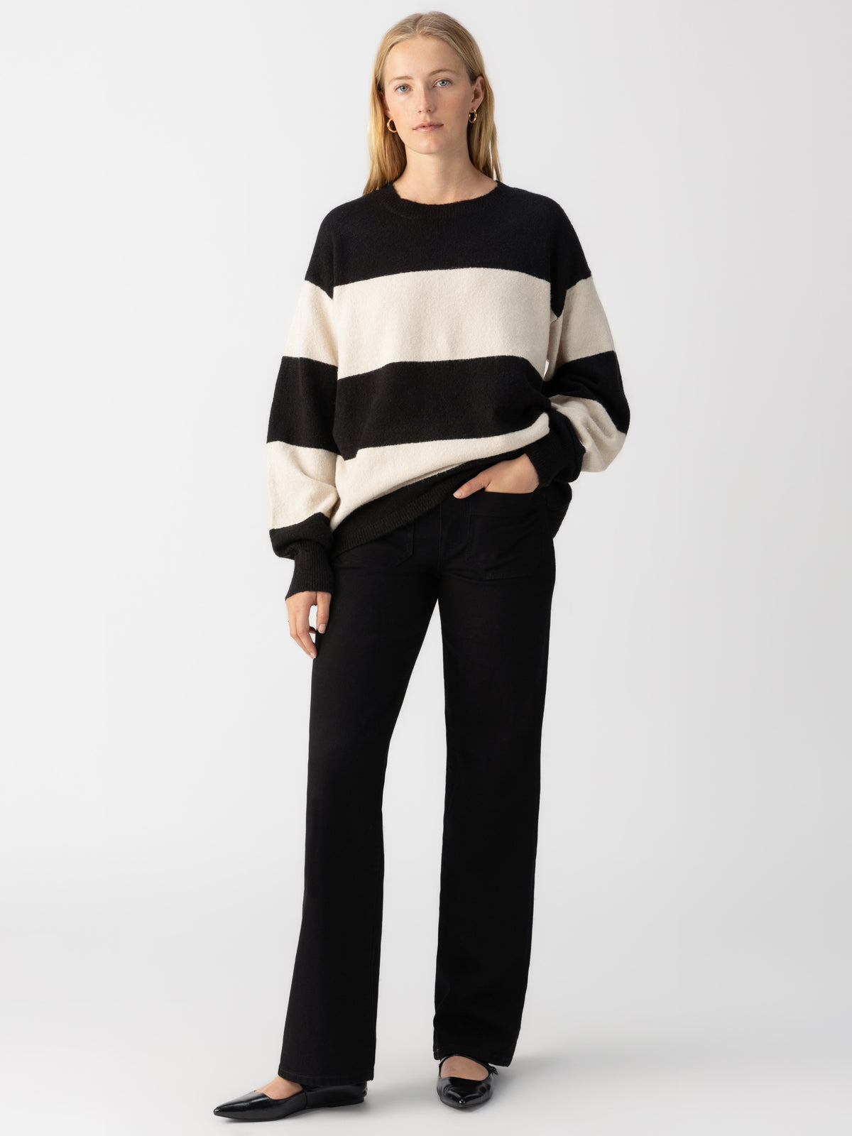 A person with long hair stands against a plain background, wearing a "Cuddle Up Crewneck Toasted Almond Black Stripe" sweater by Sanctuary Clothing, along with black pants and black shoes. Their left hand is in their pocket as they look straight ahead.