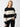 A person stands against a plain white background, wearing the Cuddle Up Crewneck Toasted Almond Black Stripe sweater from Sanctuary Clothing and black pants. The person has long blonde hair, is looking directly at the camera, and has one hand in their pocket.