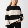 A person stands against a plain white background, wearing the Cuddle Up Crewneck Toasted Almond Black Stripe sweater from Sanctuary Clothing and black pants. The person has long blonde hair, is looking directly at the camera, and has one hand in their pocket.