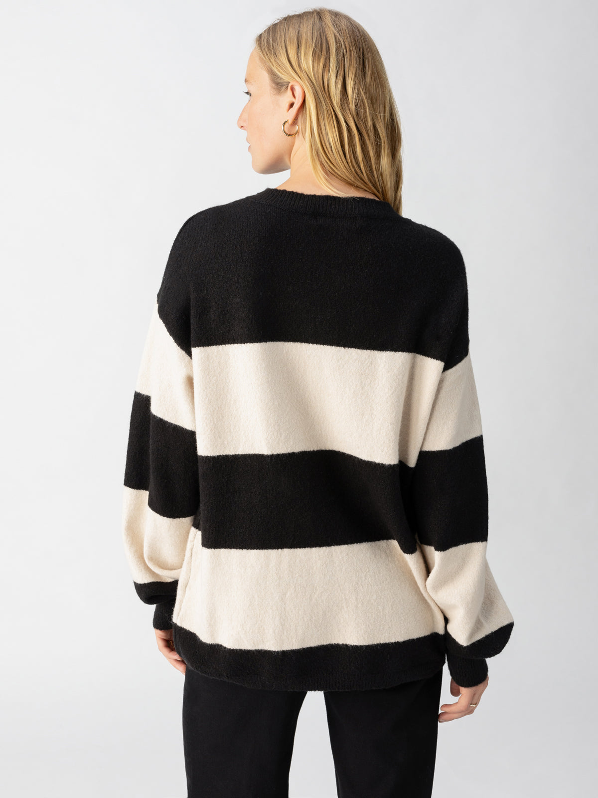 A person with long blonde hair is turned away from the camera, wearing the Sanctuary Clothing Cuddle Up Crewneck Toasted Almond Black Stripe. The sweater features a loose fit with large horizontal black and white stripes, showcased against a plain, light-colored background that highlights the outfit.