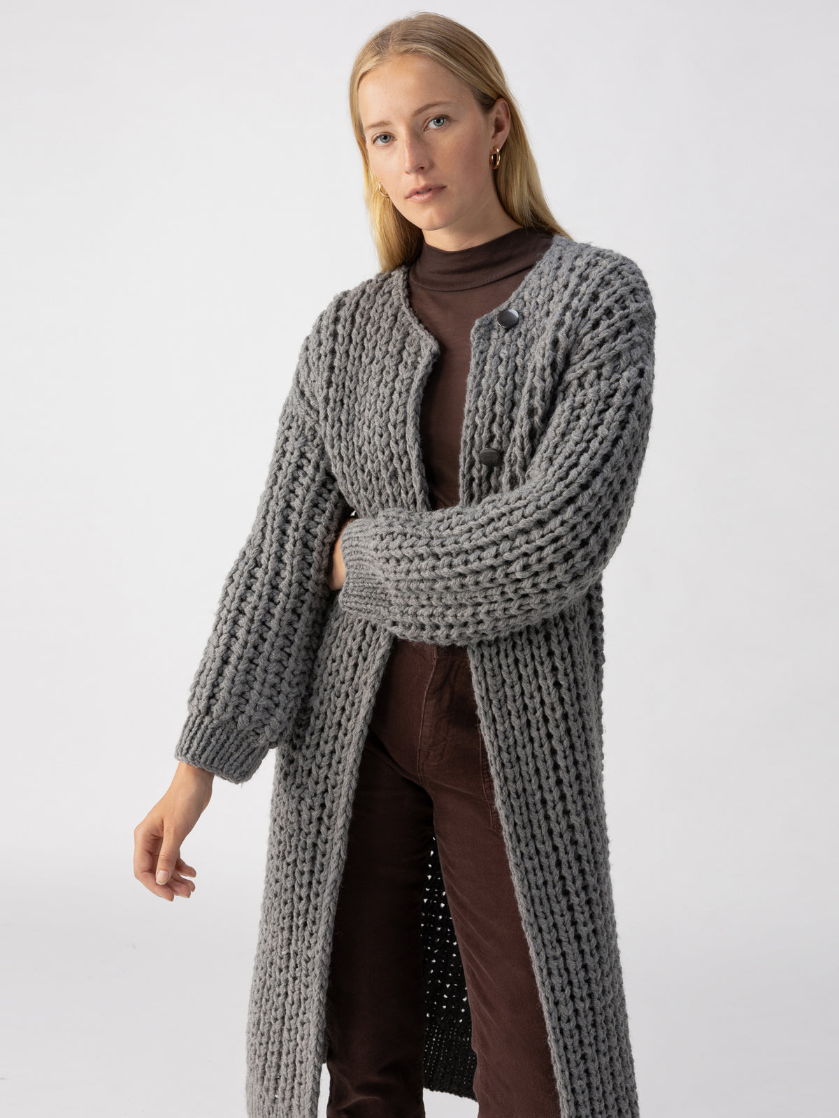 A person with long blonde hair is wearing The Ultimate Cardi Heather Ash by Sanctuary Clothing, a long, chunky, knitted grey cardigan over a brown turtleneck and brown pants. The background is plain white, and the person has a neutral expression with one arm slightly lifted.