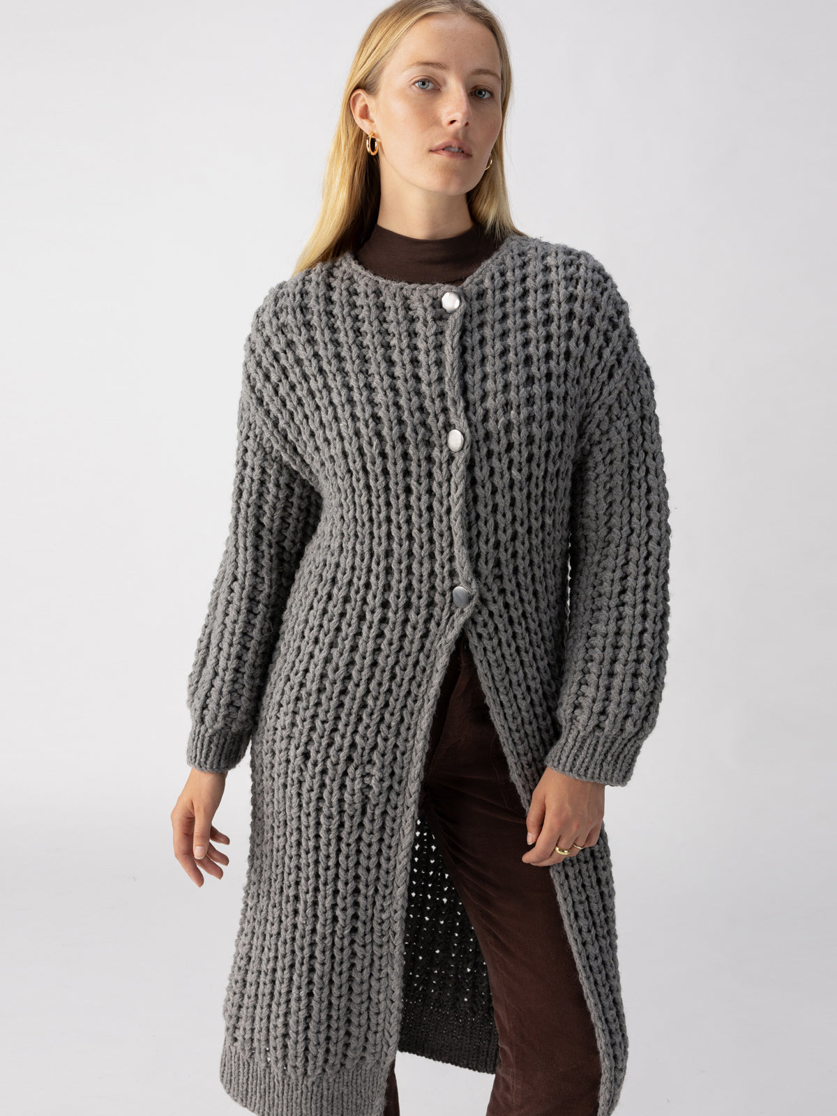 A person with long blond hair is wearing The Ultimate Cardi Heather Ash by Sanctuary Clothing, a long chunky knit gray cardigan with large buttons, over a brown top and brown pants. They are standing against a plain beige background, looking forward with a neutral expression.