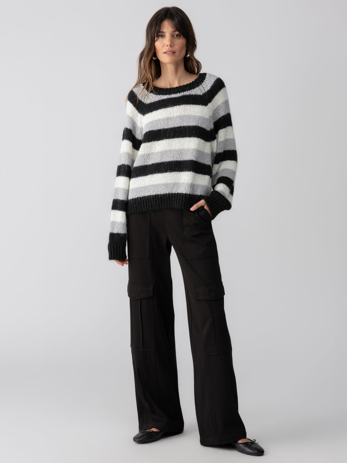 A person with long hair is wearing the Sanctuary Clothing Picture This Stripe Sweater in Ash Multi Stripe and black wide-leg pants. They have one hand in their pocket and are looking slightly to the side against a plain gray background.