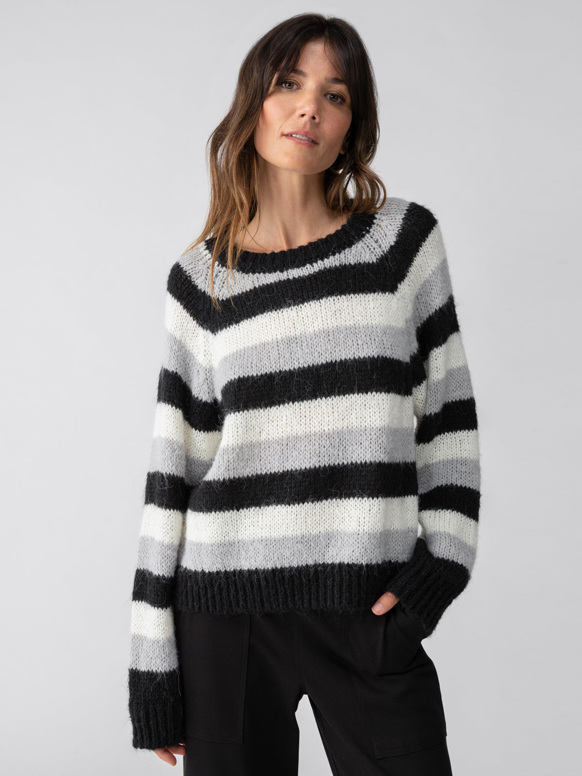 A person with long hair is wearing the Sanctuary Clothing Picture This Stripe Sweater in Ash Multi Stripe, featuring wide black, white, and gray horizontal stripes, while posing with one hand in their pocket against a plain light gray background.