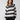 A person with long hair is wearing the Sanctuary Clothing Picture This Stripe Sweater in Ash Multi Stripe, featuring wide black, white, and gray horizontal stripes, while posing with one hand in their pocket against a plain light gray background.