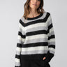 A person with long hair is wearing the Sanctuary Clothing Picture This Stripe Sweater in Ash Multi Stripe, featuring wide black, white, and gray horizontal stripes, while posing with one hand in their pocket against a plain light gray background.