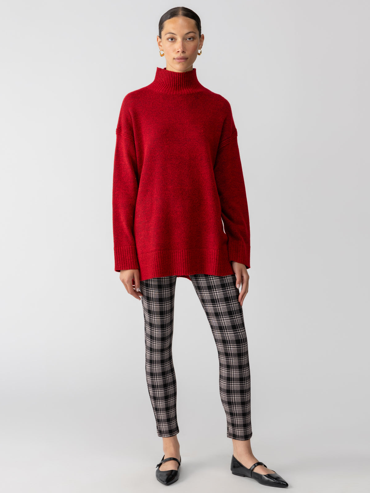 A person wearing the Sanctuary Clothing Perfect Tunic in Mars Red, plaid leggings, and black flats stands against a plain gray background. They have their hair tied back and wear dangling earrings.