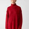 A person dressed in the Perfect Tunic Mars Red from Sanctuary Clothing and plaid pants stands against a plain white background. Their hair is pulled back, and they are adorned with hoop earrings.