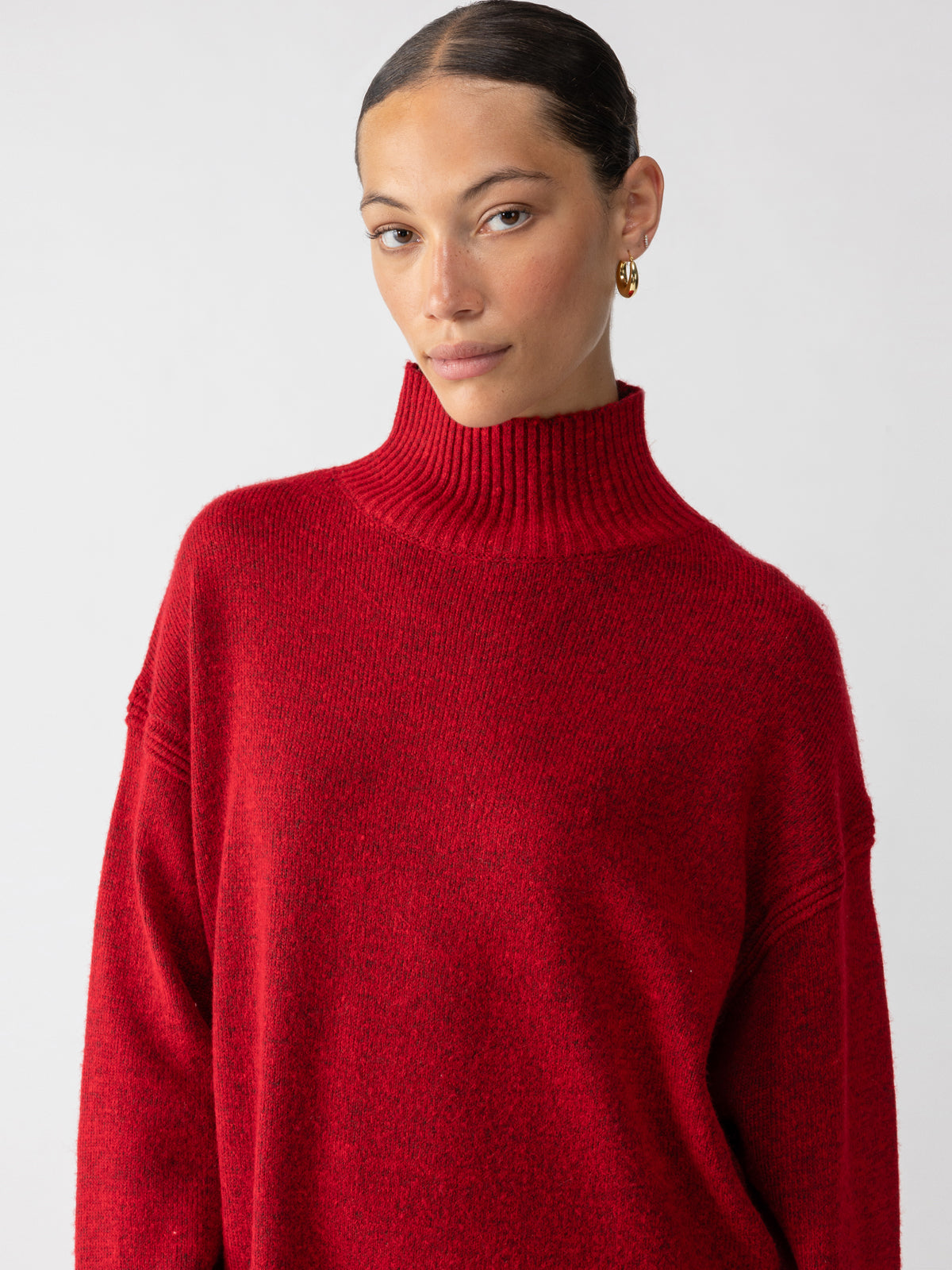 A person with tied-back hair is wearing the Perfect Tunic Mars Red by Sanctuary Clothing and gold hoop earrings, standing against a plain white background with a calm expression.