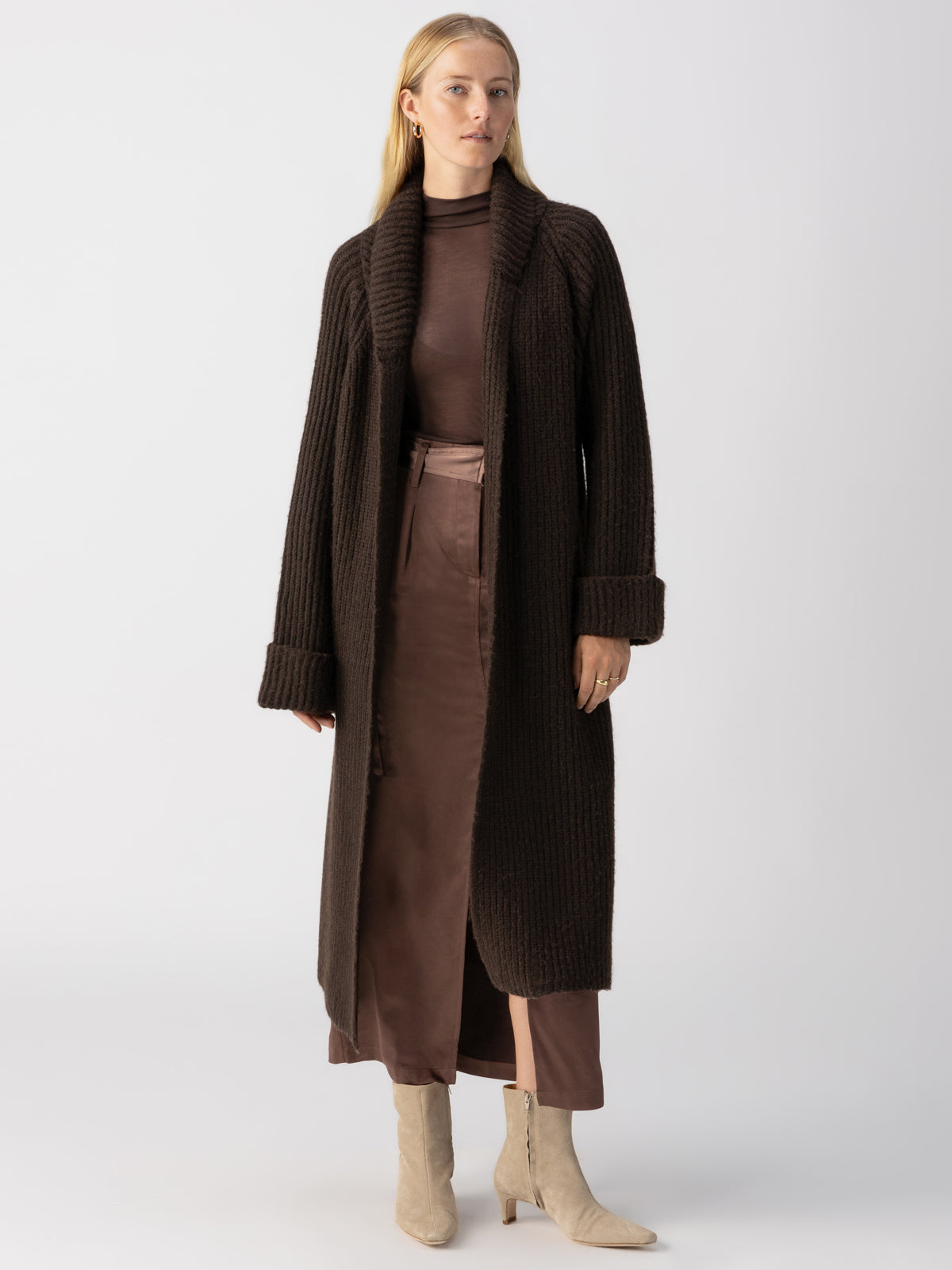 A woman with long blonde hair stands against a white background, wearing a brown, long-sleeved dress paired with the dark brown "All I Need Cardi Coffee" by Sanctuary Clothing. She completes her look with light-colored ankle boots and minimal accessories, gazing straight ahead.