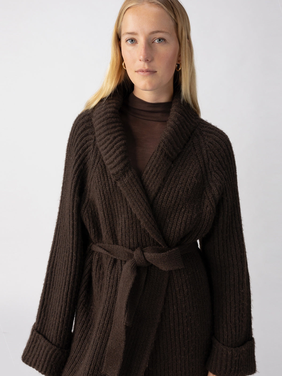 A person with long blonde hair wears the "All I Need Cardi Coffee," a dark brown knitted cardigan from Sanctuary Clothing, paired with a sheer brown turtleneck and a waist belt. They have a neutral expression and are looking directly at the camera. The background is plain and light-colored.