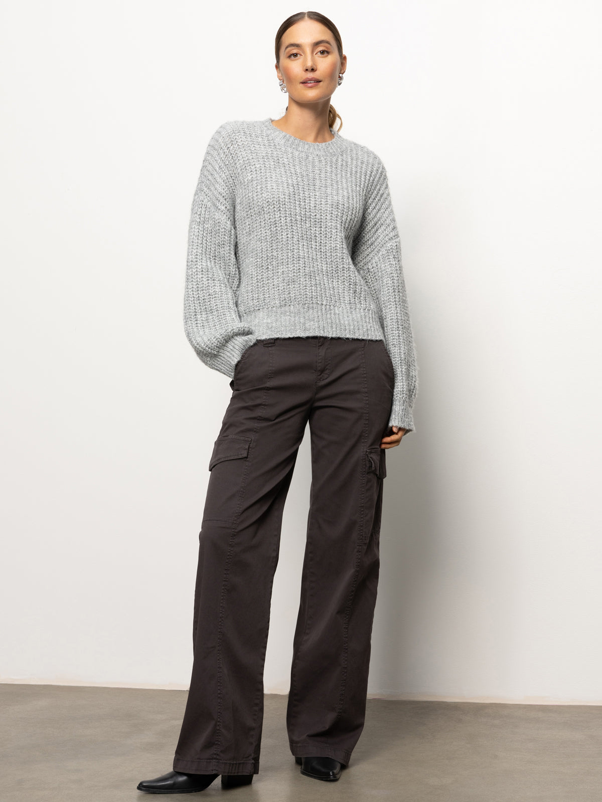 A person stands against a plain background, wearing the Sanctuary Clothing Fuzzy Volume Sleeve Sweater in Heather Grey and dark cargo pants. They have shoulder-length hair tied back and are looking confidently at the camera, with their hands relaxed by their sides.