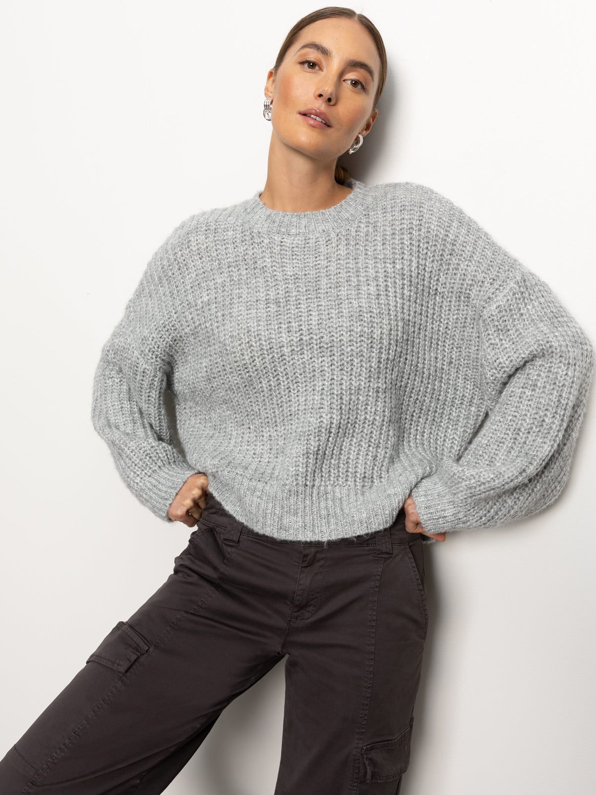 A person poses against a plain background, wearing the Fuzzy Volume Sleeve Sweater in Heather Grey from Sanctuary Clothing and dark cargo pants. Their hair is pulled back, and they look confidently at the camera.