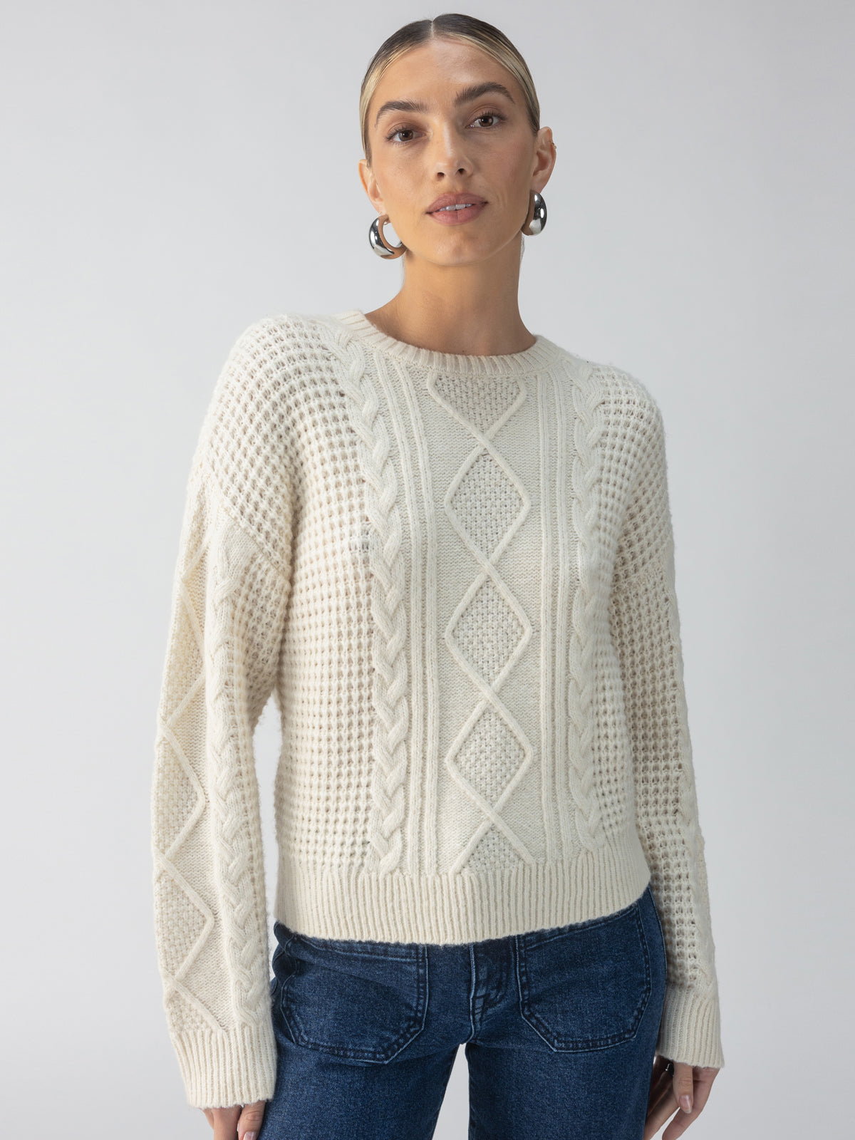 A person wearing the Sanctuary Clothing Tie Back Cable Sweater in Frosted Almond and jeans poses against a plain background. The sweater showcases textured patterns like diamonds and cables. The individual has long hair pulled back and accessorizes with hoop earrings.