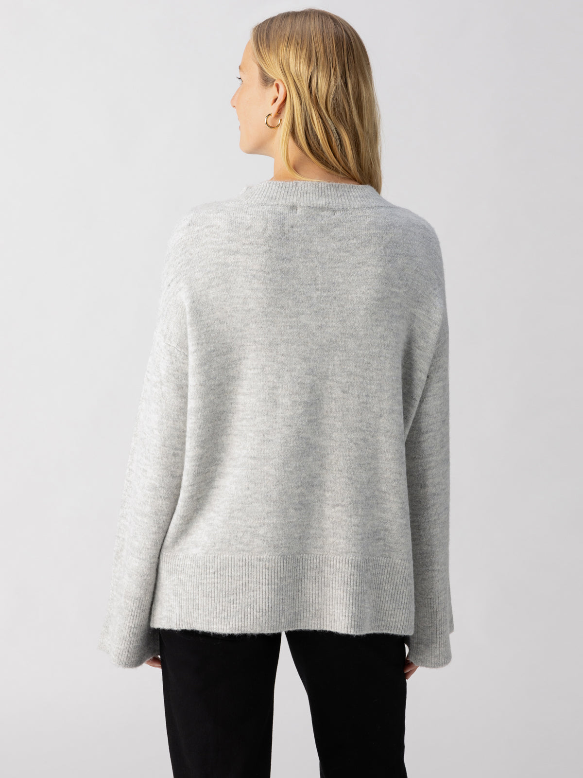 A person with long blonde hair, dressed in the Sanctuary Clothing Street Chic Sweater Heather Grey and black pants, is standing with their back turned towards the camera against a plain light gray background.