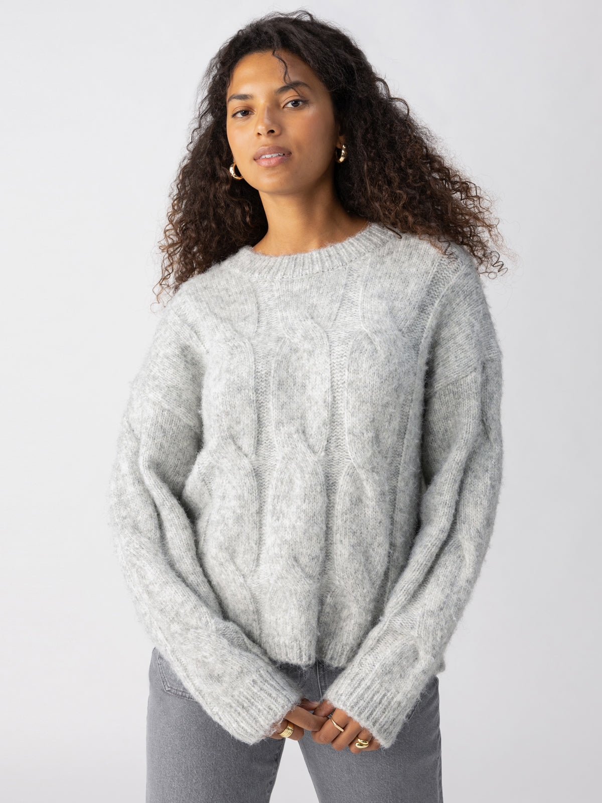 A person with curly hair is wearing a soft, heather grey Cable Crew sweater from Sanctuary Clothing along with grey pants. They are standing with their hands slightly in their pockets against a plain white background. They have a neutral expression on their face.