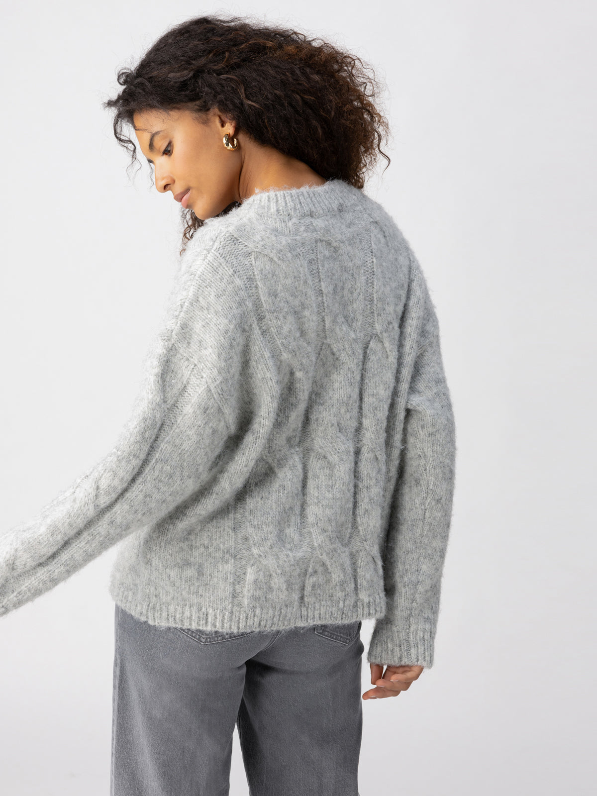 A person with long, curly hair is turned slightly to the side and looking down. They are wearing the "Cable Crew Heather Grey" sweater from Sanctuary Clothing along with grey pants. The background is plain and light-colored.