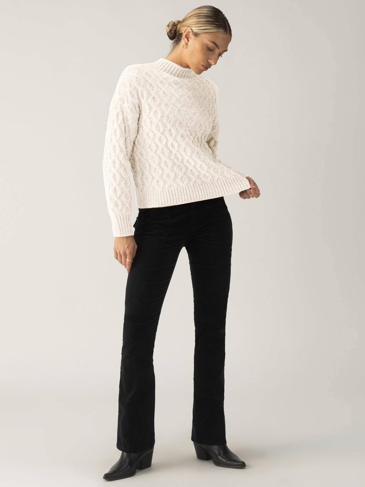 A person with tied-back hair is wearing the Sanctuary Clothing cable mock neck sweater in light almond, black pants, and dark ankle boots. They are standing against a plain background and looking down to their right.