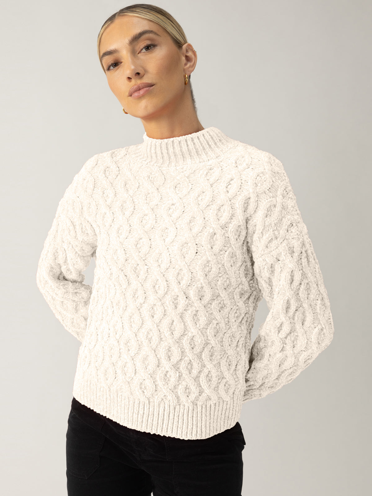 A person wearing a Sanctuary Clothing cable mock neck sweater in light almond and black pants stands with arms behind their back against a plain light background, looking confidently at the camera.