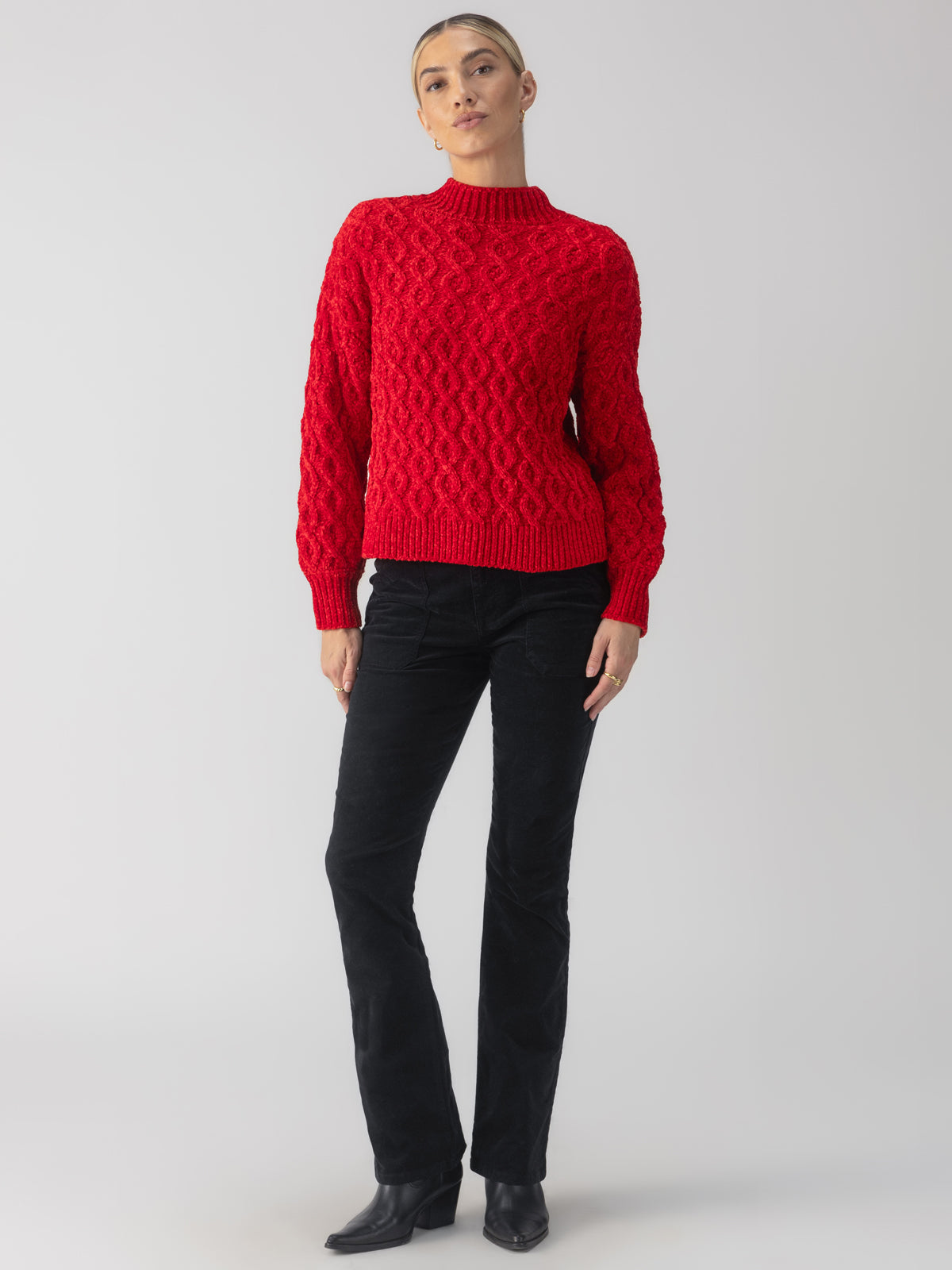 A person wearing the Cable Mock Neck Sweater Mars Red by Sanctuary Clothing and black pants stands against a plain background. They are posing confidently with a neutral expression and hands resting by their sides.